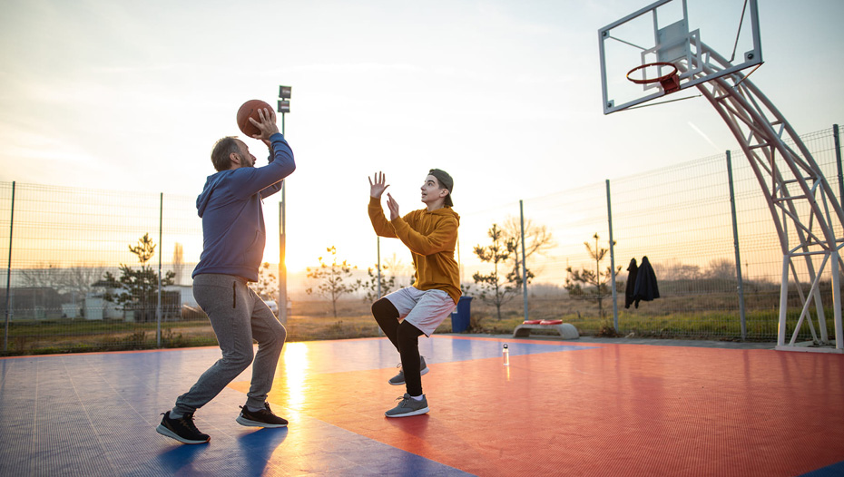 A young man and his father play basketball together on an outdoor court.